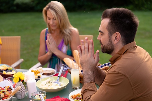 Widok z boku białej pary siedzącej na zewnątrz przy stole obiadowym nastawionym na rodzinny posiłek, z zamkniętymi oczami i rękami w modlitwie, odmawiając łaskę przed jedzeniem