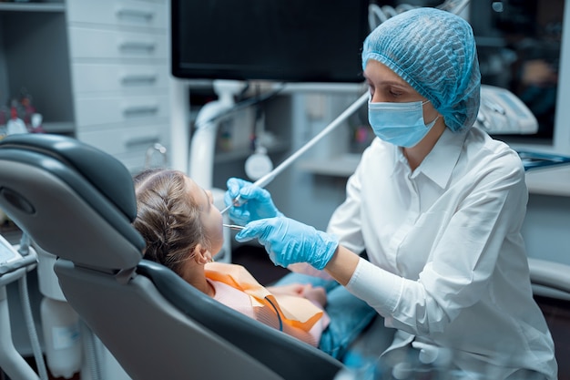 Widok z boku dentysty w mundurze medycznym, który leczy zęby małej dziewczynki pacjenta w gabinecie stomatologicznym