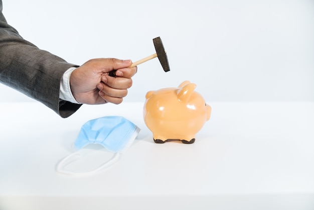 Widok z boku dłoni z młotkiem rozbijającej ceramiczną skarbonkę z białym tłem i bardzo dobrym oświetleniem oraz maską na twarz w związku z pandemią koronawirusa COVID19
