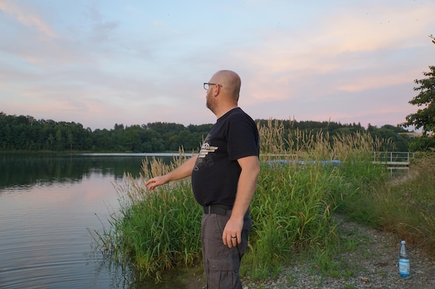 Widok z boku dojrzałego mężczyzny stojącego na brzegu jeziora na tle nieba podczas zachodu słońca