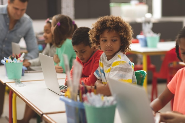 Widok z boku dzieci w wieku szkolnym korzystających z laptopa w klasie, podczas gdy nauczyciel uczy w tle