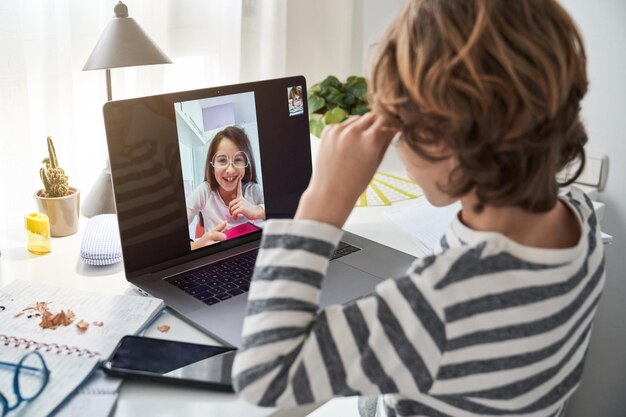 Widok z boku dziecka rozmawiającego na laptopie z dziewczyną w okularach, siedzącego przy stole ze smartfonem w pokoju