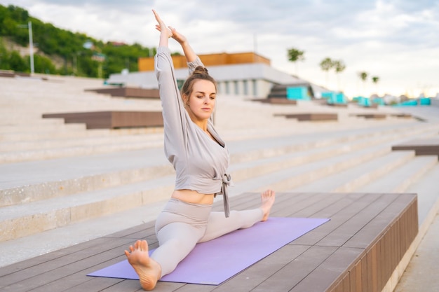 Widok z boku elastycznej szczupłej sportowej kobiety noszącej odzież sportową siedzącą na sznurku z rękami na macie do jogi na zewnątrz