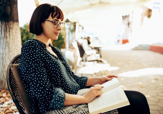Widok z boku inteligentnej młodej kobiety w zielonej koszuli w białe kropki i przezroczyste okulary siedzącej na zewnątrz na ulicy miasta i czytającej książkę Młoda studentka ucząca się na ulicy miasta