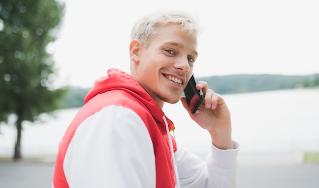 Widok z boku kaukaski przystojny blond hipster mężczyzna uśmiechający się podczas rozmowy na telefonie komórkowym, siedząc w parku Ludzie komunikacji podróży i koncepcji nowoczesnych technologii