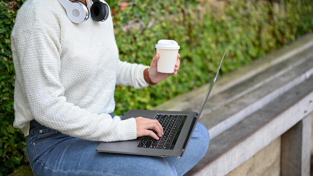 Widok z boku kobiety piszącej na laptopie, trzymając filiżankę kawy i siedząc w parku