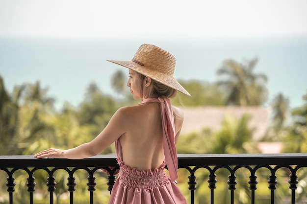 Widok z boku kobiety w różowej sukience i słomkowym kapeluszu stojącej na hotelowym balkonie, widok na ocean.