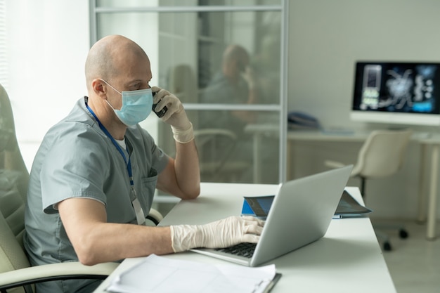 Widok z boku łysego dojrzałego klinicysty płci męskiej w masce ochronnej, siedzącego przy biurku przed laptopem w gabinecie lekarskim i rozmawiającego przez telefon komórkowy