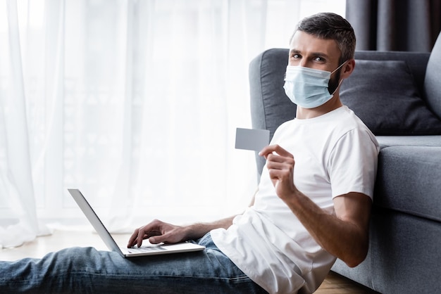 Widok z boku mężczyzny w masce medycznej patrzącego na kamerę, trzymającego kartę kredytową i używającego laptopa w