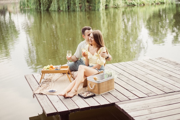 Widok z boku młodej szczęśliwej pary na romantyczny piknik w pobliżu rzeki lub jeziora, kobieta i mężczyzna razem piją wino na zewnątrz, ludzie bawią się na letnie wakacje