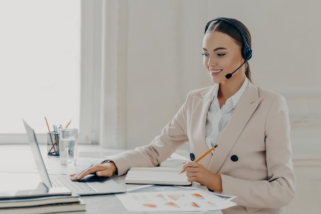 Widok z boku młodej uśmiechniętej kobiety biznesu, która bierze udział w konferencji internetowej, patrząc na operatora call center na ekranie laptopa, konsultując się z klientem, siedząc za biurkiem w nowoczesnym biurze