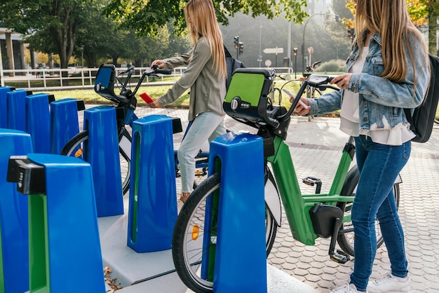 Widok z boku na dwie kobiety używające smartfona do wypożyczenia dwóch rowerów z wypożyczalni rowerów, aby jeździć po mieście.