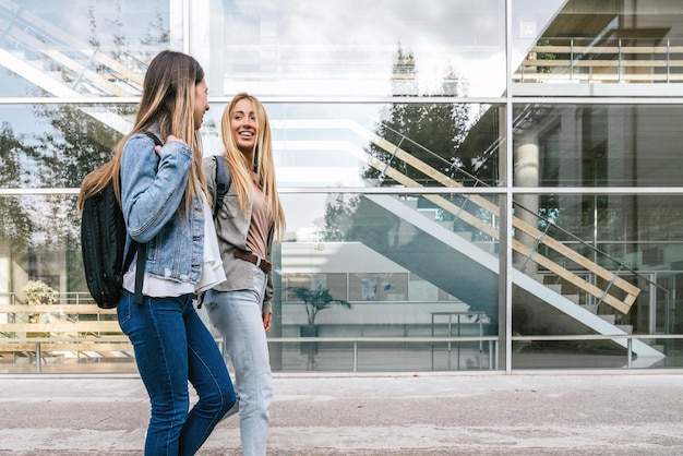 Widok z boku na dwie koleżanki z college'u spacerujące podczas rozmowy ze szklanym budynkiem w tle.