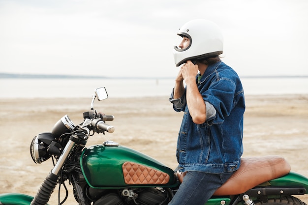 Widok z boku na przystojnego młodego mężczyznę w swobodnym stroju, siedzącego na motocyklu na plaży, w kasku