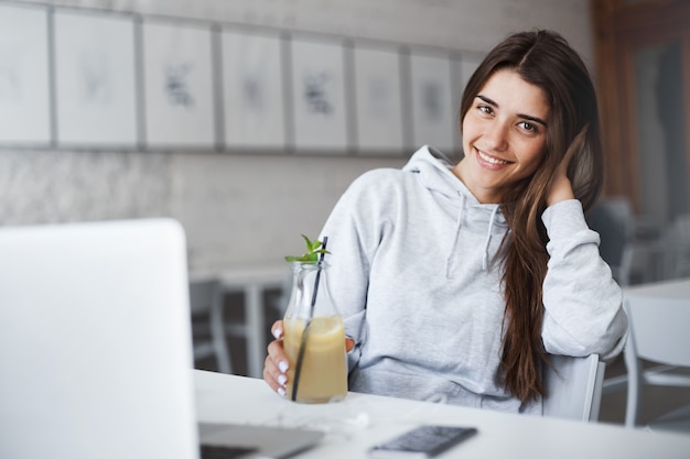 Widok Z Boku Portret Atrakcyjnej Brunetki W Kawiarni, Opierając Rękę Na Krześle, Uśmiechając Się I Trzymając Drinka, Siedząc Przed Smartfonem I Laptopem.