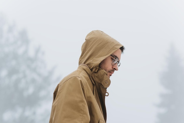Widok z boku portret mężczyzny słuchającego muzyki za pomocą słuchawek i telefonu komórkowego, patrząc w dół i noszący kaptur płaszcza na okularach głowy spaceru w mglistym parku Koncepcja ludzi i technologii