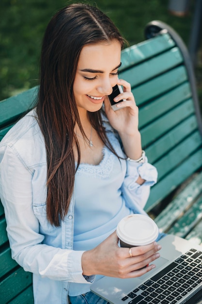 Widok Z Boku Portret Piękny Młody Student Siedzi Na Plaży W Pobliżu Uniwersytetu, Picia Kawy I Korzystania Z Laptopa Podczas Rozmowy Przez Telefon Z Uśmiechem.