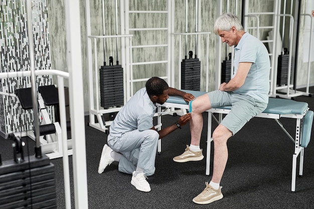 Widok z boku portret specjalisty rehabilitacji zakładającego taśmę na kolano starszego pacjenta w fizjocie