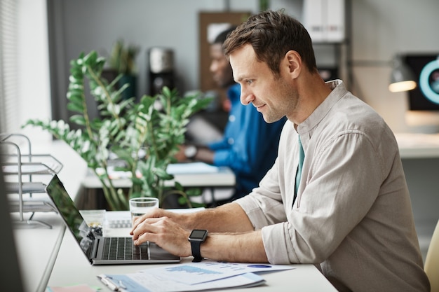 Widok z boku portret uśmiechniętego dorosłego mężczyzny korzystającego z laptopa podczas pracy w przestrzeni biurowej