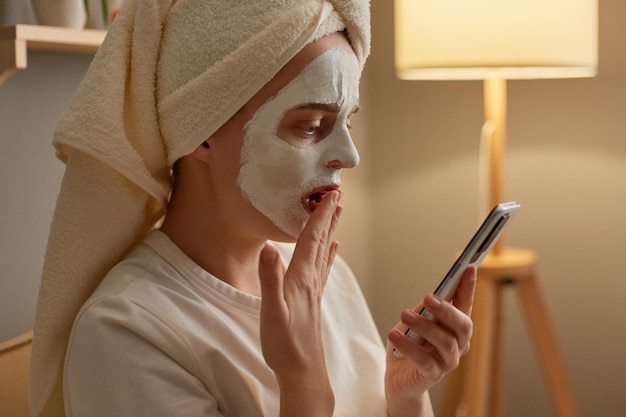 Widok z boku portret zdumionej kaukaskiej kobiety z peelingującą maską na twarzy w białym ręczniku, siedzącej na kanapie w domu, wykonującej zabiegi kosmetyczne, trzymając smartfon, patrząc na wyświetlacz zakrywający usta dłonią