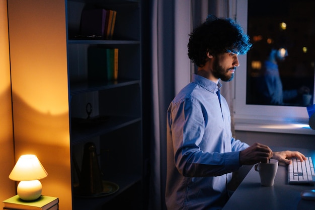 Widok z boku poważnego młodego biznesmena pracującego na komputerze w ciemnym biurze domowym w nocy