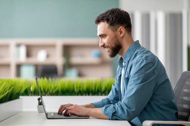 Widok z boku przystojnego młodego mężczyzny korzystającego z laptopa w nowoczesnym biurze