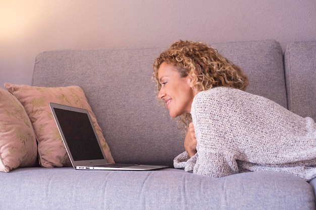 Widok z boku radosnej, uśmiechniętej, szczęśliwej dorosłej kobiety korzystającej z laptopa leżącego na kanapie w domu podczas relaksu w pomieszczeniach sama Kobieta surfująca po sieci nwe Technologia bezprzewodowe połączenie Nowoczesna