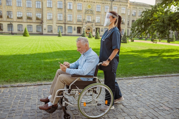 Widok z boku starszego mężczyzny odzyskującego pacjenta na wózku inwalidzkim za pomocą smartfona na spacerze z pielęgniarką