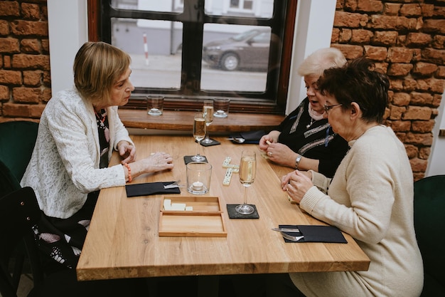 Widok z boku starszych ciemnowłosych i jasnowłosych kobiet w czarno-białych kurtkach, grających w gry planszowe w kawiarni z przyjaciółmi świętujących spotkanie przy stole z kieliszkami wina lub szampana na nim.