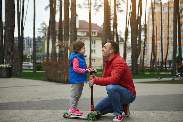 Widok z boku uroczej dziewczynki na hulajnodze, uśmiechającej się radośnie, patrząc na swojego ojca stojącego z nim twarzą w twarz w parku miejskim w piękny słoneczny jesienny dzień