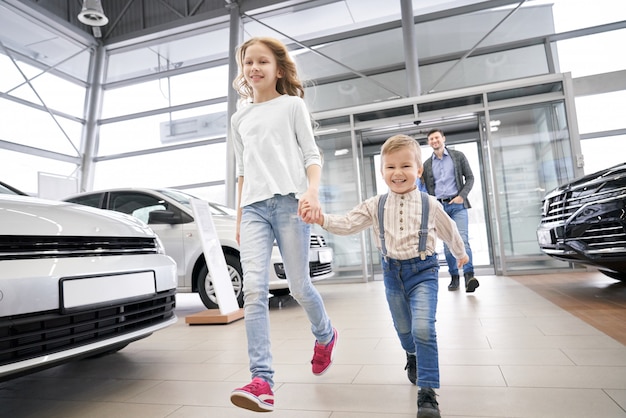 Widok z boku uśmiechniętego dwojga dzieci idących w dużym salonie samochodowym