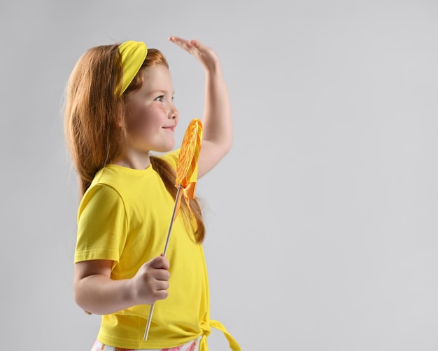 Widok z boku uśmiechniętej małej rudowłosej dziewczynki w letnim modnym stroju, trzymającej lizaki i witającej kogoś ręką w górę Strzał studio