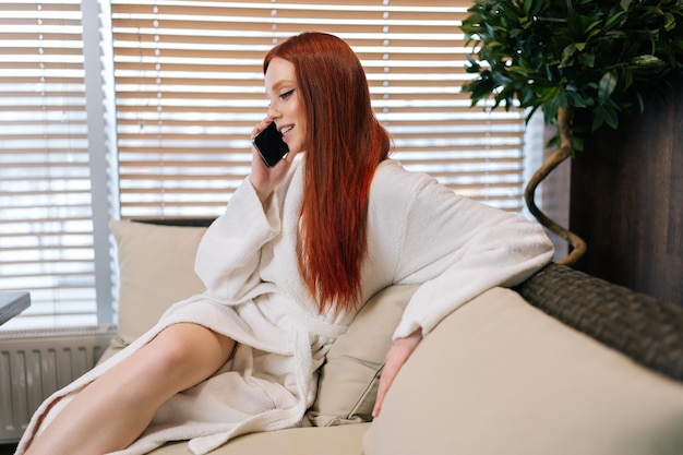 Widok z boku uśmiechniętej młodej kobiety w białym szlafroku siedzącej na wygodnej sofie przy oknie i rozmawiającej na smartfonie, cieszącej się rozmową po zabiegach spa w salonie piękności
