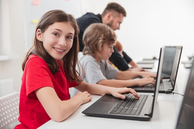 Widok z boku uśmiechniętej nastoletniej uczennicy patrzącej w kamerę podczas korzystania z laptopa podczas lekcji z kolegami z klasy i nauczycielem w nowoczesnej szkole