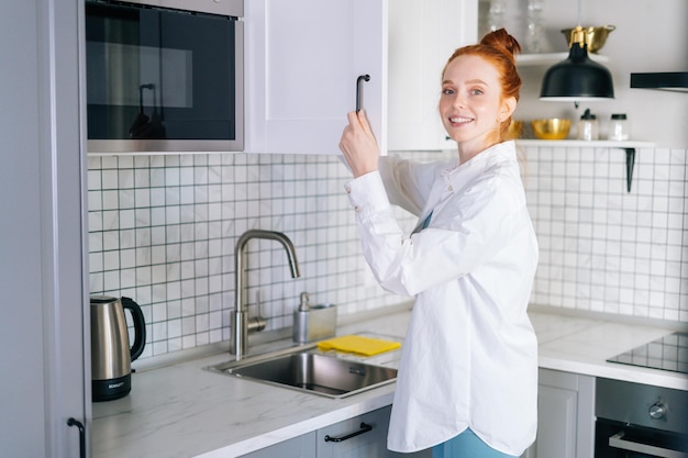 Widok z boku uśmiechniętej rudej młodej kobiety otwierającej drzwi szafki kuchennej w jasnym pokoju kuchennym