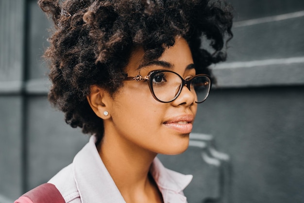 Widok z boku zbliżenie młoda african american studentka w okularach szykuje się do pójścia na studia pozowanie na ulicy Piękna kobieta nastolatek afro z plecakiem pozowanie na szarej ścianie odkryty