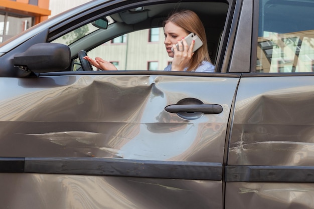 Widok z boku zdenerwowanej kobiety o blond włosach, siedzącej w rozbitym samochodzie i rozmawiającej przez telefon, dzwoniącej do agenta ubezpieczeniowego lub męża, ma problemy z zadrapaniami i wgnieceniami drzwi samochodu Zdjęcie na zewnątrz