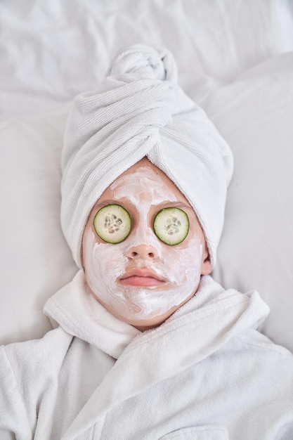 Widok z góry chłopca z maską na twarz i plasterkami ogórka, ubranego w turban z ręcznikiem i szlafrok, relaksującego się na miękkim łóżku podczas zabiegów kosmetycznych w sypialni w domu