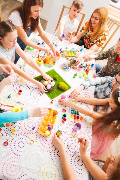 Widok z góry dzieci dekorujących kolorowe jajka trzymające się za ręce na stole. Grupa dzieci malujących tradycyjne pisanki na dużym stole z białym obrusem