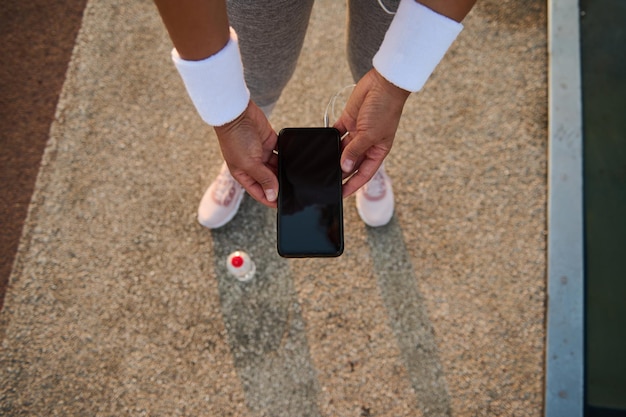 Widok z góry kobiece lekkoatletka ręce w białe opaski frotte, trzymając smartfon z pustym pustym czarnym ekranem i przeglądanie stron internetowych i aplikacji mobilnych.