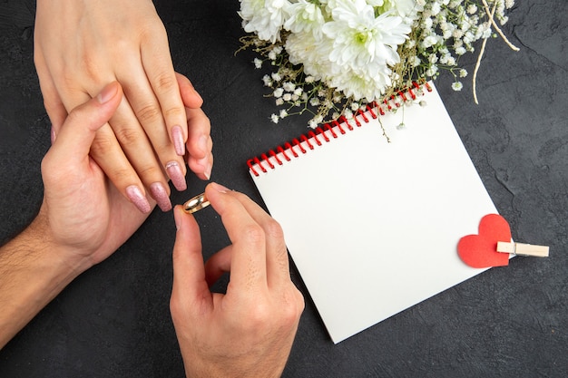 widok z góry koncepcja propozycji małżeństwa męskie dłonie umieszczające pierścionek na kobiecej dłoni kwitnie notatnik na ciemnym tle