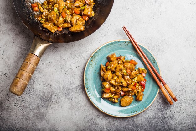 Widok z góry kurczaka Kung Pao na talerzu gotowym do spożycia. Smażone chińskie danie tradycyjne z kurczakiem, orzeszkami ziemnymi, warzywami, papryczkami chili. Chiński obiad, pałeczki, rustykalne betonowe tło