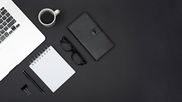 Widok z góry laptopa; herbata; długopis; notes spiralny; okulary do oczu; pamiętnik i spinacz do papieru na czarnym tle