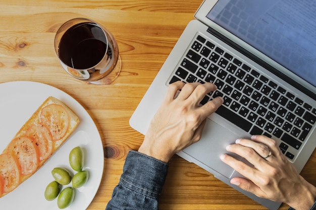 Widok z góry męskich rąk do pracy z laptopem podczas lekkiego wieczornego chleba, oliwek i wina. Płaskie świeckich koncepcja pracy na komputerze podczas kolacji w kuchni