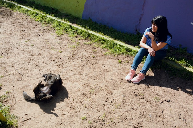 widok z góry młoda kobieta siedzi w parku i obserwuje, jak jej pies tarza się na ziemi