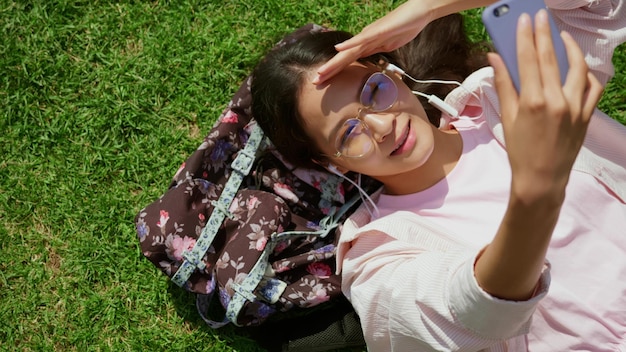 Widok z góry młodej azjatyckiej kobiety leżącej na trawie na zewnątrz po rozmowie wideo ze słuchawkami i telefonem komórkowym