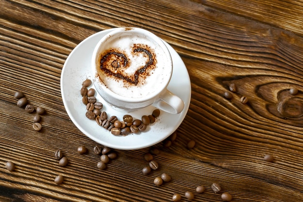 Widok z góry na ceramiczną filiżankę gorącej kawy cappuccino latte z rysunkiem w kształcie serca z cynamonem lub kakao z szablonem na piance mlecznej, ziarnach kawy na drewnianym stole w kawiarni, z bliska