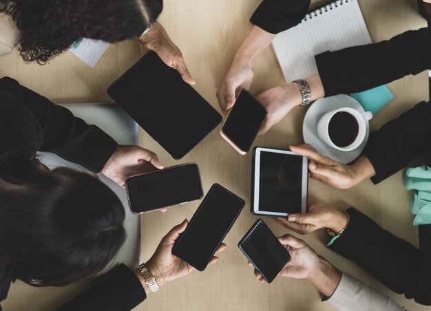 Widok z góry na drewniany stół konferencyjny z sześcioma dłońmi kobiet biznesu trzymających smartfony i tablety wokół stołu. Koncepcja spotkania biznesowego.