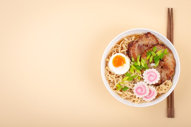 Widok z góry na japońską zupę z makaronem ramen w białej misce z makaronem, bulionem mięsnym, plastrami pieczonej wieprzowiny, narutomaki, jajkiem z żółtkiem na pastelowym beżowym tle. Tradycyjne danie japońskie, miejsce na tekst