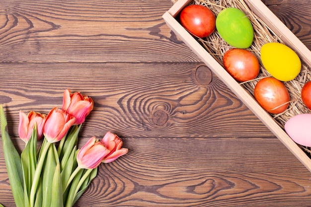 Widok z góry na kolorowe jajka wielkanocne na łóżku ze słomy w długim drewnianym pudełku na drewnianym stole i czerwone tulipany na tekst